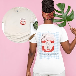 May include: A cream-colored t-shirt with a small red and white jam jar graphic on the front. The back of the shirt features a larger graphic of a jam jar with the text "JESUS IS MY JAM" and a Bible verse on a blue gingham background.