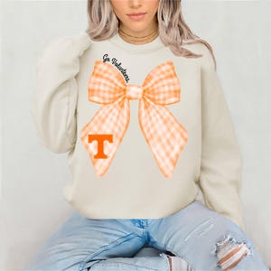 May include: A cream-colored sweatshirt with a large orange and white gingham bow and the text "Go Volunteers" above the bow. The bow has a large orange "T" in the center.