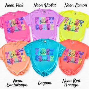 May include: A collection of neon-colored t-shirts with the words "First Grade" printed on the front in a colorful, block letter design. The shirts are in shades of pink, violet, lemon, cantaloupe, lagoon, and red-orange.