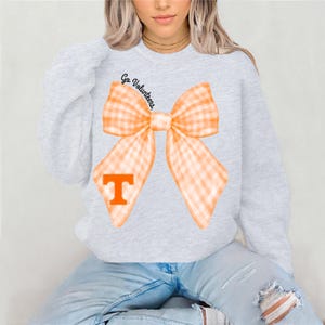 May include: Grey sweatshirt with a large orange and white gingham bow and the text "Go Volunteers." A large orange "T" is in the centre of the bow.