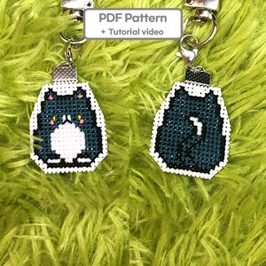 May include: Two keychains with a cross-stitch cat design. The cat is dark blue with a white belly and face, and a white outline. The keychains have silver-colored hardware. The image also includes the text "PDF Pattern + Tutorial video".