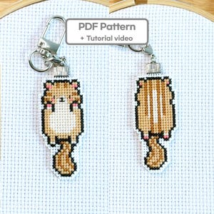 May include: Two cross-stitch keychains featuring a cat design. One shows the front of the cat, the other the back. The keychains are tan, brown, and pink, with silver-colored hardware. The text "PDF Pattern + Tutorial video" is in the background.