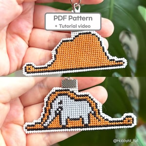 May include: A handmade keychain featuring two designs: a hat and an elephant. The hat is orange with a black outline and white border. The elephant is gray with an orange outline and white border. Text on the image reads "PDF Pattern + Tutorial video."