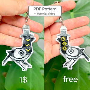 May include: A cross-stitch keychain featuring a pigeon design with a top hat. The bird is primarily black and white with yellow accents. The keychain has a silver-colored clasp and the text "PDF Pattern + Tutorial video" is displayed above the keychain.