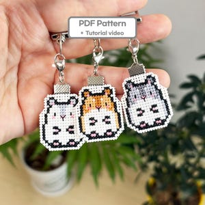 May include: Three cross-stitch hamster keyrings in white, orange, and grey. Each keyring has a silver clasp and a small, rectangular metal piece at the top. The keyrings are held together by a silver clasp. The text "PDF Pattern + Tutorial video" is in a white rectangle.