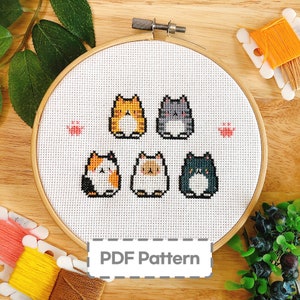 Peut inclure: Un motif de point de croix représentant cinq adorables chats de dessin animé dans différentes couleurs, notamment orange, noir, blanc et gris. Le motif est affiché sur un tissu blanc tendu sur un cerceau à broder en bois. Le texte "PDF Pattern" est visible en bas de l'image.