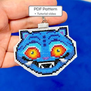 May include: A blue cross-stitch keychain featuring a tiger face with orange and yellow eyes, white fangs, and a white border. The keychain has a silver metal clasp and the text "PDF Pattern + Tutorial video" in a white box.