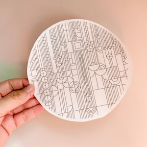 May include: A circular, off-white coaster with a black geometric design. The design features lines, circles, and rectangular shapes arranged in an abstract pattern. The coaster is held against a light pink background.