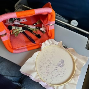 May include: An open orange and pink striped tote bag filled with embroidery floss and pens. A wooden embroidery hoop holds a piece of fabric with a cactus and butterfly design. The scene suggests a crafting or DIY project.