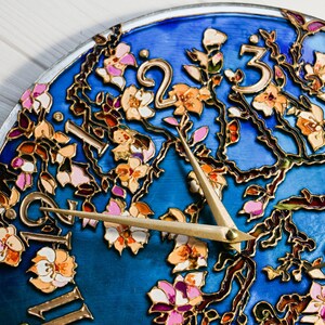 May include: A blue clock face with gold painted branches and pink and white flowers. The clock hands are gold and point to the numbers 2 and 3.