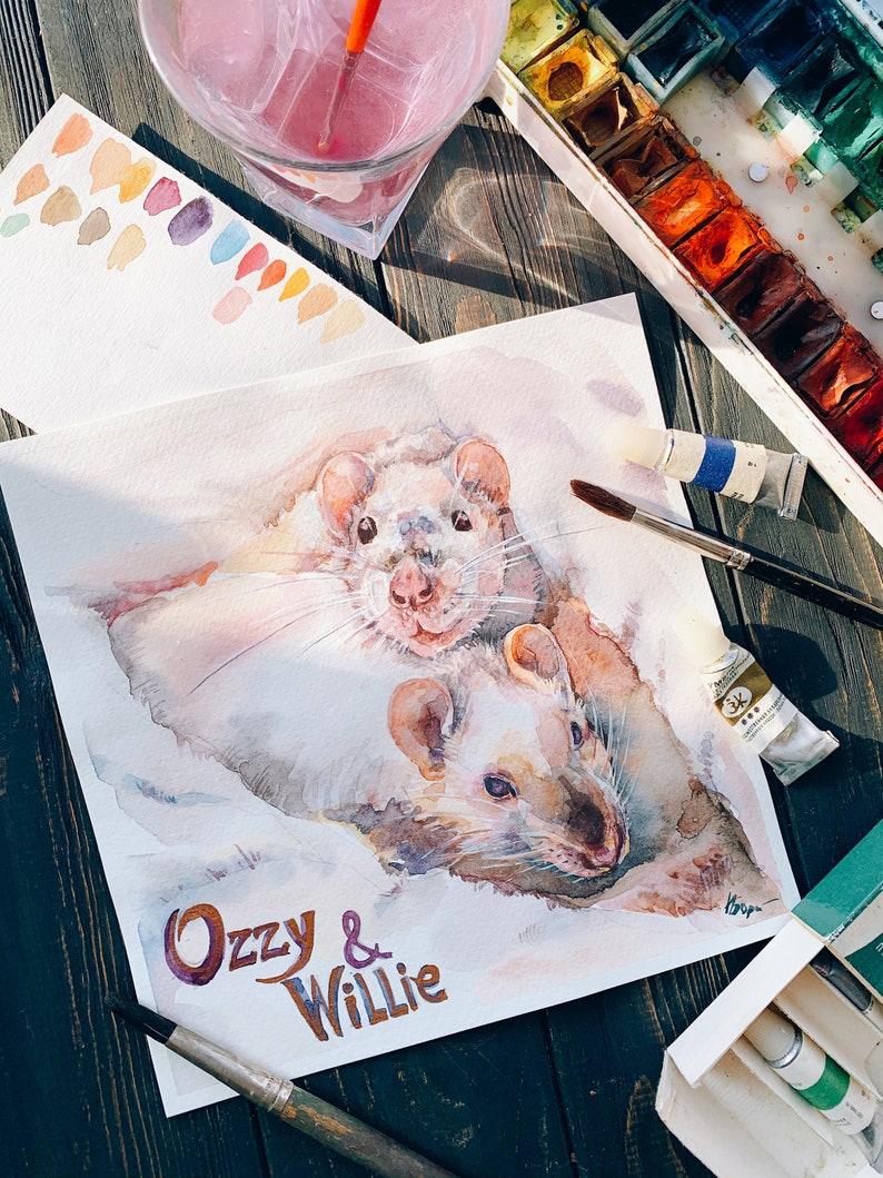 May include: Watercolor painting of two white rats, titled "Ozzy & Willie." The artwork features soft, blended colors and is surrounded by watercolor supplies, including paint tubes, brushes, and a palette of colors. The painting is on a white paper.