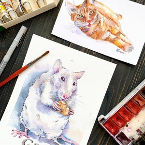 May include: Watercolor paintings of a ginger cat and a white rat, with the word "GRAY" below the rat. The cat is lying down, and the rat is holding a piece of food. Paint tubes, a paintbrush, and a watercolor palette are also visible.