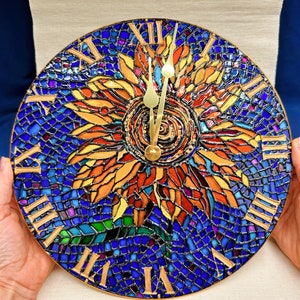 Puede incluir: Un reloj redondo con un diseño de mosaico que presenta un girasol en tonos de naranja, amarillo y rojo. La esfera del reloj es azul con marcas de números romanos.
