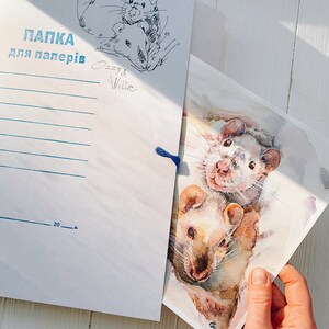May include: A white paper folder with blue text and a sketch of two rats, labeled "Ozzy & Willie." A watercolor painting of two rats is held in front of the folder. The painting has a watercolor effect with shades of brown and white.