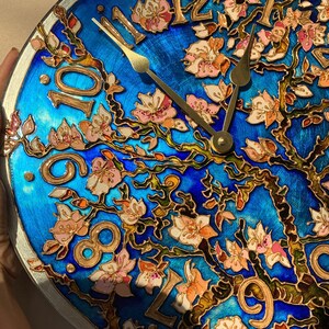 May include: A blue and gold clock with a floral design. The clock face features Roman numerals and gold hands. The floral design is painted in shades of pink, white, and green.