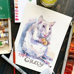 May include: Watercolor painting of a white rat holding a piece of food. The rat is named "Gray" in the painting. The painting is surrounded by art supplies, including paint tubes, a palette, and a glass of lemonade.