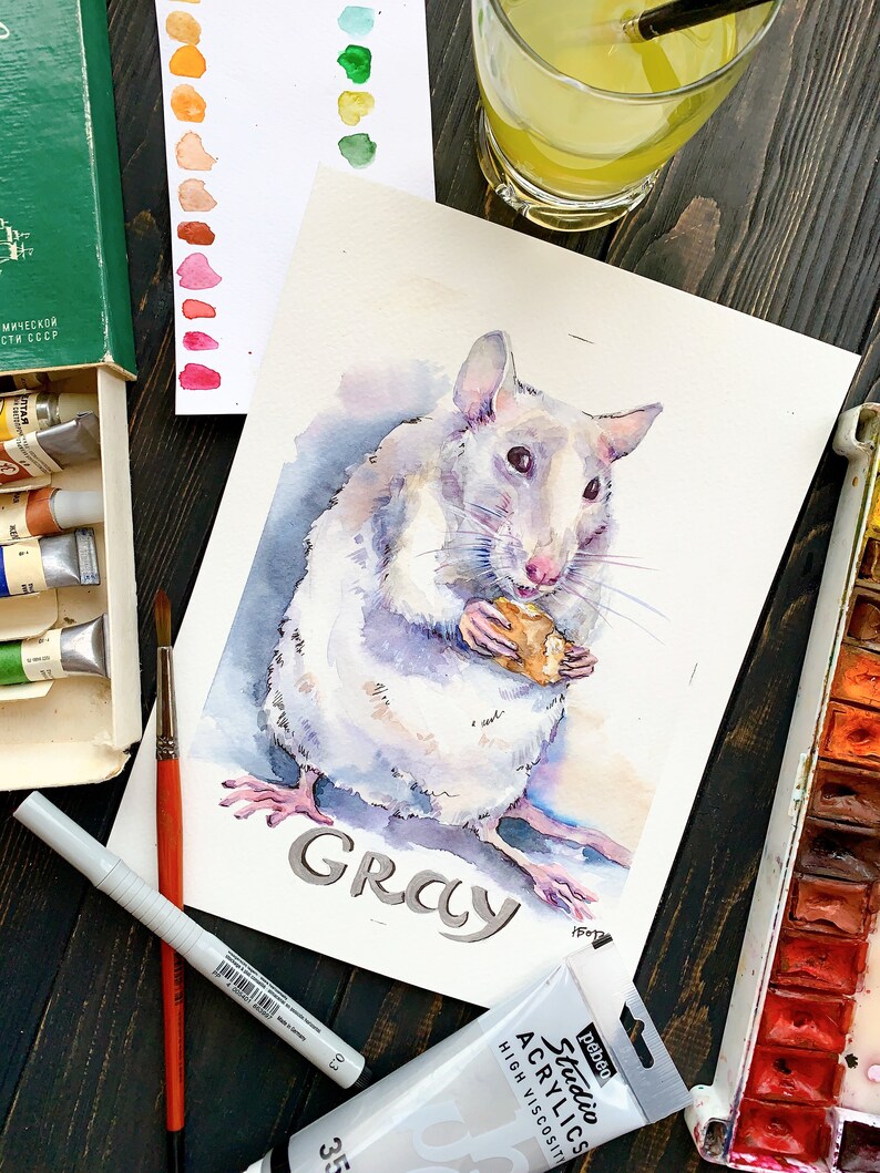 May include: Watercolor painting of a white rat holding a piece of food. The rat is named "Gray" in the painting. The painting is surrounded by art supplies, including paint tubes, a palette, and a glass of lemonade.