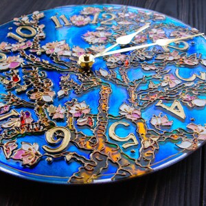 May include: A blue and gold clock with a tree design and pink flowers. The clock face is made of glass and has Roman numerals.