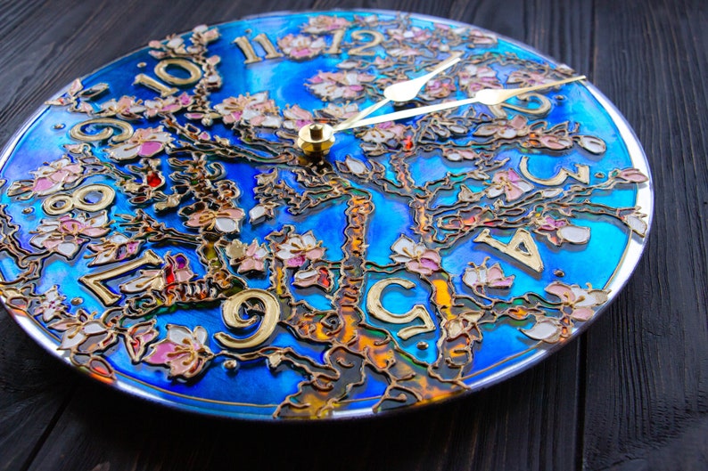 May include: A blue and gold clock with a tree design and pink flowers. The clock face is made of glass and has Roman numerals.