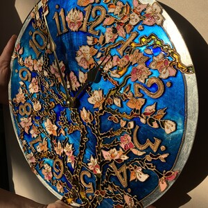 May include: A round clock with a blue background and a gold tree with pink flowers. The clock face has Roman numerals and gold hands.