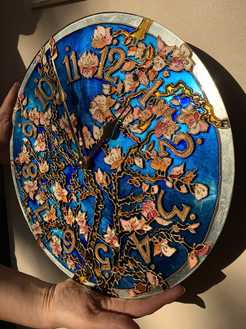 May include: A round clock with a blue background and a gold tree with pink flowers. The clock face has Roman numerals and gold hands.