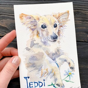 May include: Watercolor painting of a light-colored dog named Teddi, holding a small blue and white flower. The dog has long ears and a fluffy coat. The name Teddi is written in blue script. The artist's signature is in the bottom right corner.