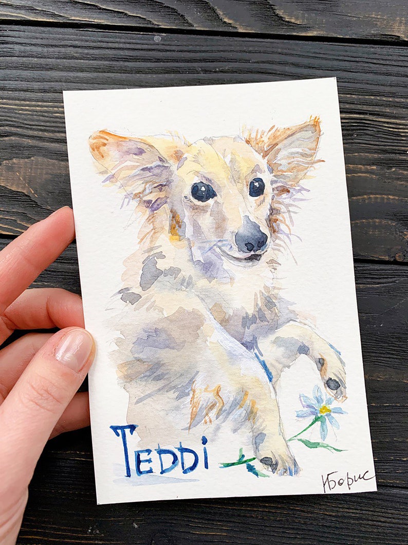 May include: Watercolor painting of a light-colored dog named Teddi, holding a small blue and white flower. The dog has long ears and a fluffy coat. The name Teddi is written in blue script. The artist's signature is in the bottom right corner.