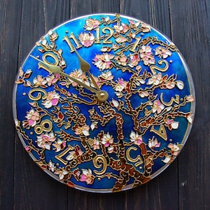 May include: A blue circular clock with gold Roman numerals and a gold tree with pink flowers. The clock is on a dark wood background.