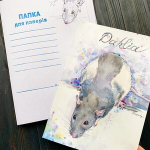 May include: Watercolor painting of a rat named Dahlia, with gray and white fur, and a pink nose. The painting is on a white paper, with colorful paint splatters. A matching notebook with the name "Dahlia" and the text "ПАПКА для паперів" is also visible.