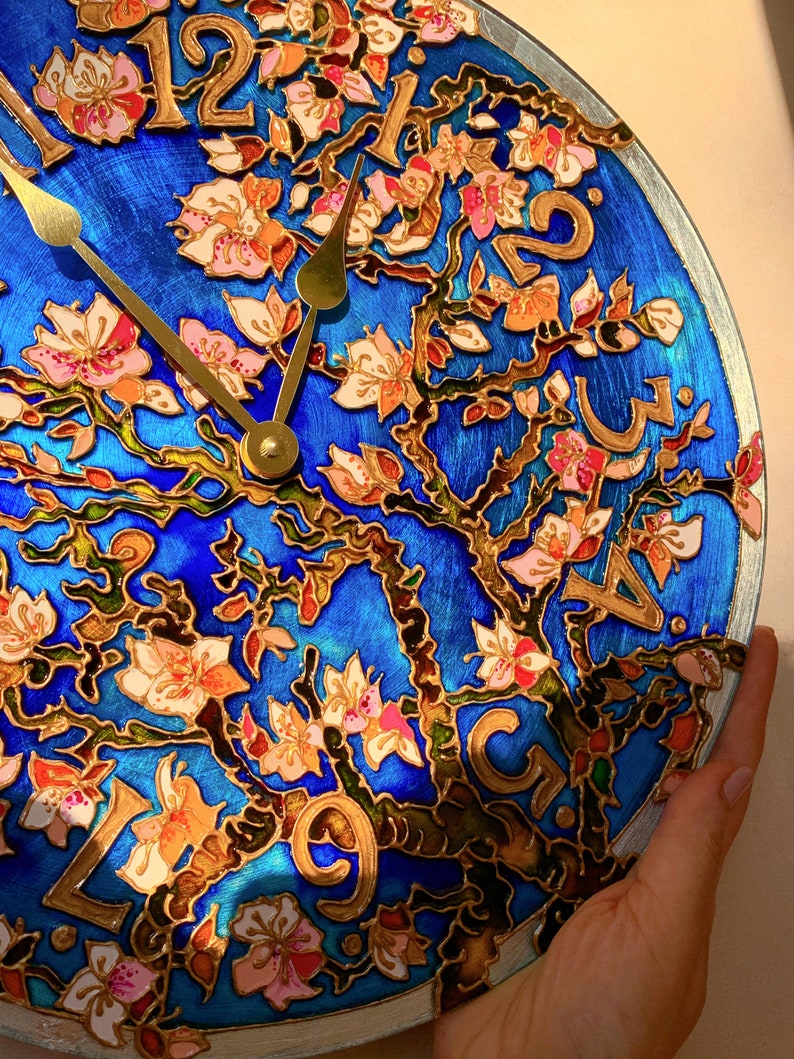 May include: A blue and gold clock with a floral design. The clock face is decorated with a tree with pink and white flowers. The numbers are in gold and the hands are gold.