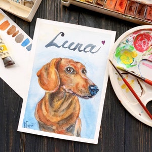 May include: Watercolor painting of a dachshund dog with brown fur and a blue nose. The name "Luna" is written above the dog in cursive script, with a small purple heart. The painting is on white paper.