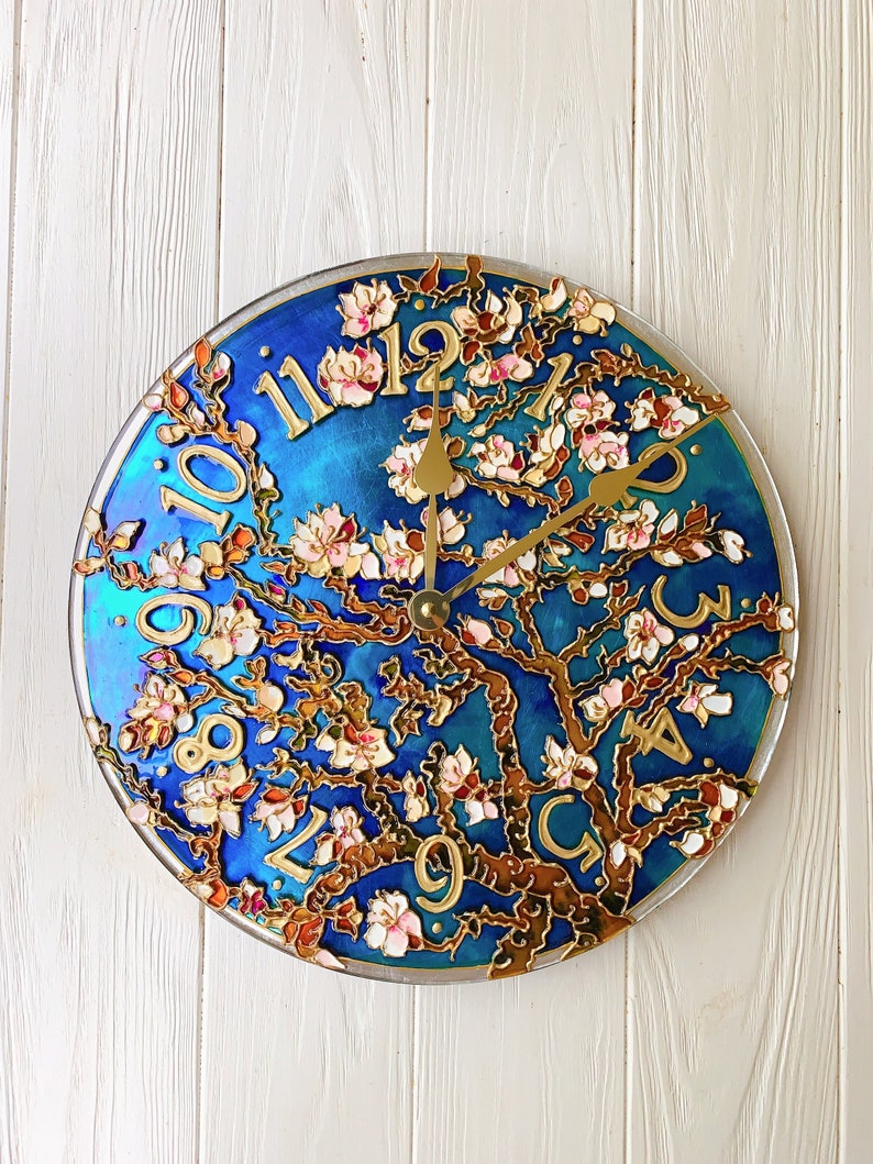 May include: A blue and gold clock with a painted design of a blooming tree. The clock face has Roman numerals and a gold-colored hour and minute hand.