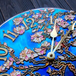 May include: A blue clock face with gold painted floral details and gold Roman numeral hour markers. The clock hands are gold and white.