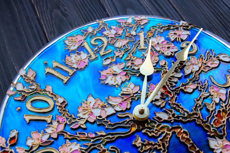 May include: A blue clock face with gold painted floral details and gold Roman numeral hour markers. The clock hands are gold and white.