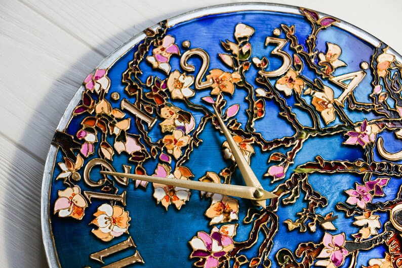 May include: A blue clock face with gold painted branches and pink and white flowers. The clock hands are gold and point to the numbers 2 and 3.