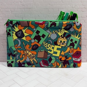 May include: A rectangular pencil case with a green zipper, filled with pens. The case features a vibrant, repeating pattern of Minecraft characters and elements in shades of green, orange, and brown. The design includes pixelated figures and TNT blocks.