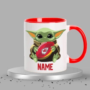 May include: White ceramic mug with a red rim and handle. The mug features a cartoon image of a green alien holding a football with the Kansas City Chiefs logo. The text "NAME" is printed below the image.