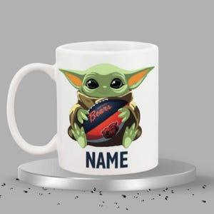 May include: White ceramic mug featuring a green cartoon character holding a football with the text "Bears". The mug also has the text "NAME" in navy blue. The character has large black eyes and pointed ears.