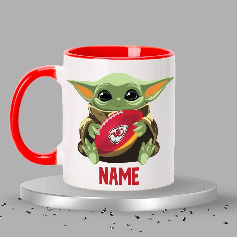 May include: A white ceramic mug with a red rim and handle. The mug features a cartoon image of a green baby Yoda holding a red and white football with the Kansas City Chiefs logo. The text "NAME" is printed below the image.