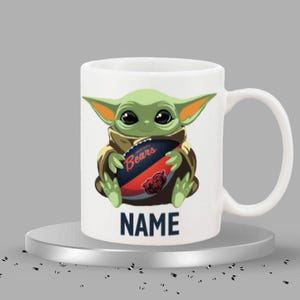 May include: White ceramic mug featuring a green alien character holding a football with the Chicago Bears logo. The mug has the word "NAME" printed in navy blue at the bottom.