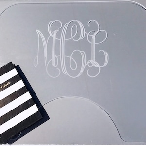 Op de afbeelding: Een grijze acryl bureaumat met een wit monogram van de letters "MGC" in een schrijflettertype. Een zwart-wit gestreept notitieboekje ligt op de mat.