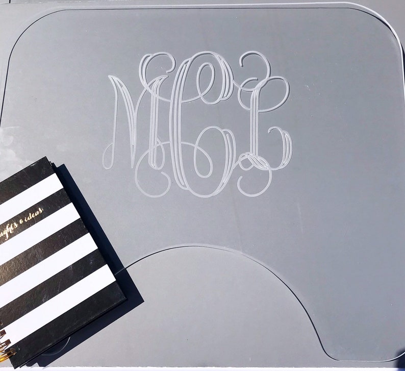 Op de afbeelding: Een grijze acryl bureaumat met een wit monogram van de letters "MGC" in een schrijflettertype. Een zwart-wit gestreept notitieboekje ligt op de mat.
