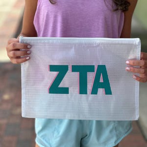 May include: White mesh zippered pouch with the letters "ZTA" in teal with pink outlines.