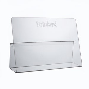 May include: Clear acrylic countertop display stand with the word "Drinkard" etched on the front. The stand has a shelf for holding items.