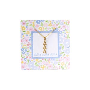 May include: A gold-colored necklace with three triangle pendants is displayed on a floral patterned card. The card has a blue border with the text "delta delta delta" printed in a blue font. The background of the card is white with colorful flowers.