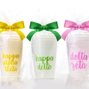 May include: Three clear plastic cups with sorority names in gold, green, and pink lettering. Each cup is wrapped in clear cellophane and tied with a matching colored bow. The cups are for a sorority event.