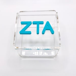Pu&ograve; includere: Una scatola in acrilico trasparente con le lettere "ZTA" in blu.