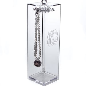 May include: Clear acrylic jewelry organizer with a rotating top and a single necklace hanging from it. The organizer has a monogrammed inscription of "E&B" on the front.