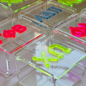 Pu&ograve; includere: Un set di scatole in acrilico trasparente con lettere color neon che compongono le parole "XO", "SB", "AM", "KA" e "XO".