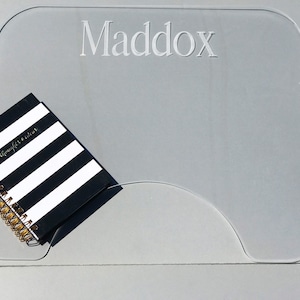 Op de afbeelding: Een transparante acryl bureaumat met de naam "Maddox" erop gegraveerd. Een zwart-wit gestreept notitieboek met een gouden spiraalbinding ligt op de mat.